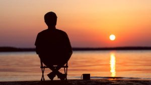 Man sits on a camping chair with a mug of tea and contemplates the beautiful sunset. The background of the sea is blurred.