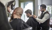 Young male barber perfecting his customer's haircut, wearing smart clothes, in a busy shop.